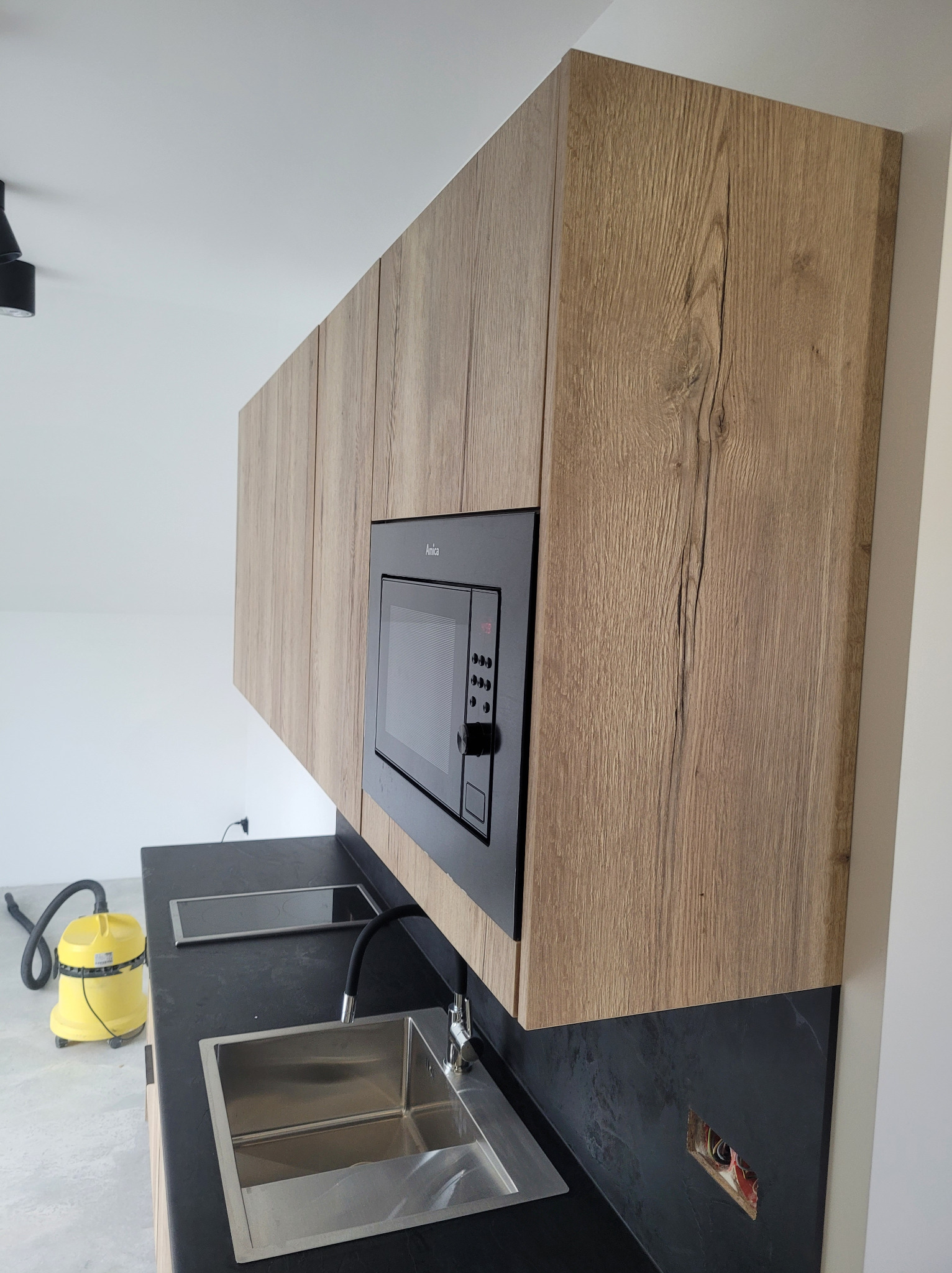 Kuchenne szafki wiszące z drewnianym dekorem i czarną mikrofalówką Amica. Na blacie zlewozmywak i płyta indukcyjna. W tle żółty odkurzacz.