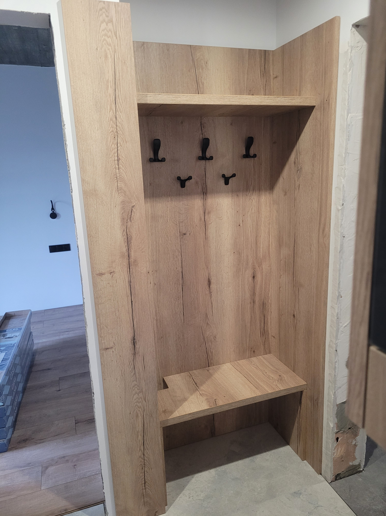 Wnękowa szafa z siedziskiem i wieszakami, wykończona drewnopodobną płytą w ciepłym odcieniu, w surowym wnętrzu przedpokoju.