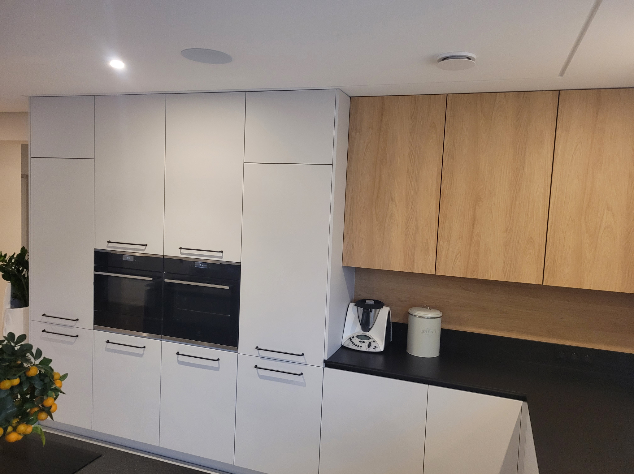 Nowoczesna kuchnia z białymi frontami i drewnianymi szafkami wiszącymi. Dwa piekarniki w zabudowie, czarny blat i minimalistyczne uchwyty. Elementy dekoracyjne.