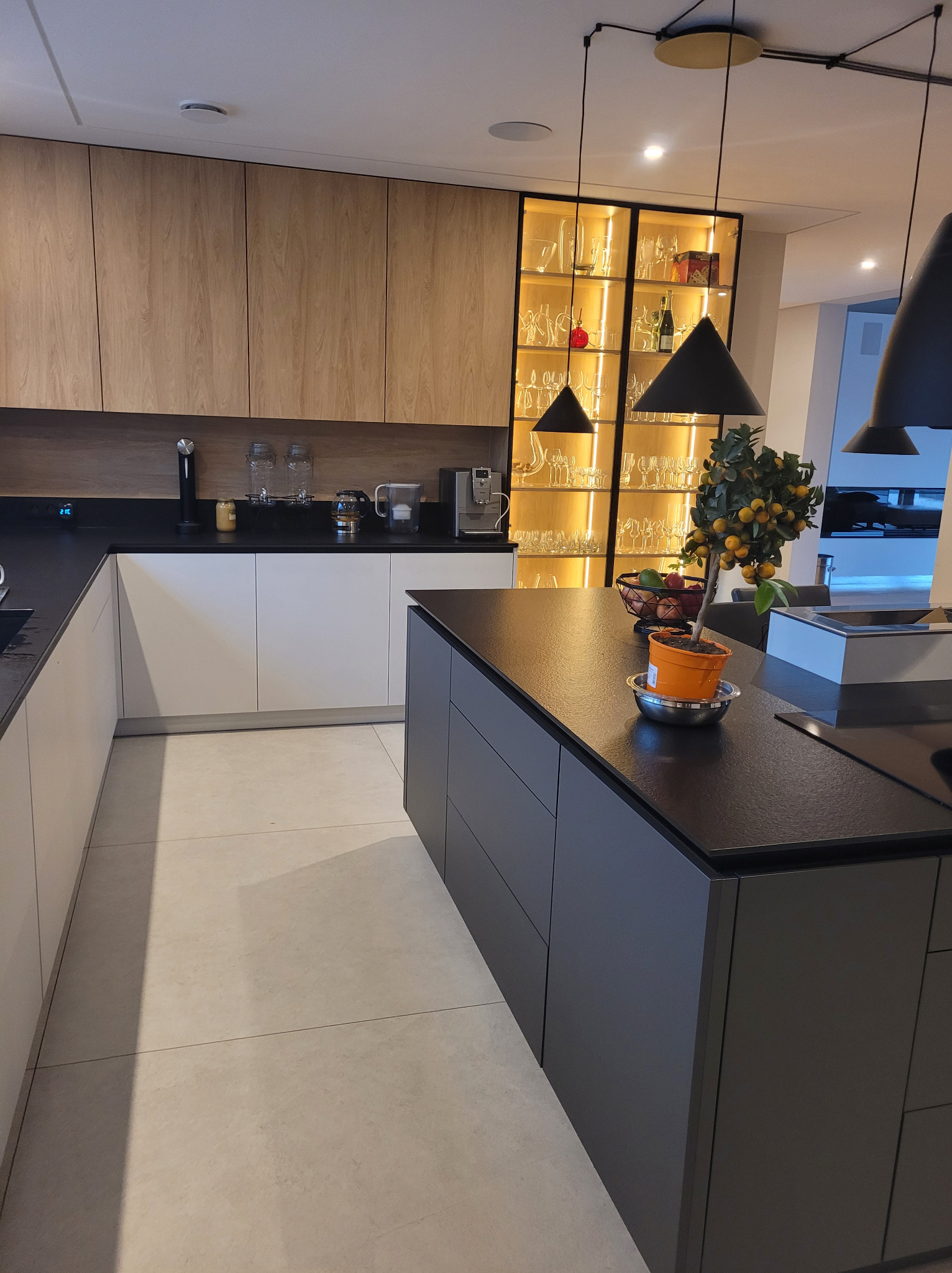 Nowoczesna kuchnia: drewniane szafki górne, białe dolne, czarny blat i wyspa. Podświetlana witryna z szkłem. Minimalistyczne lampy wiszące.