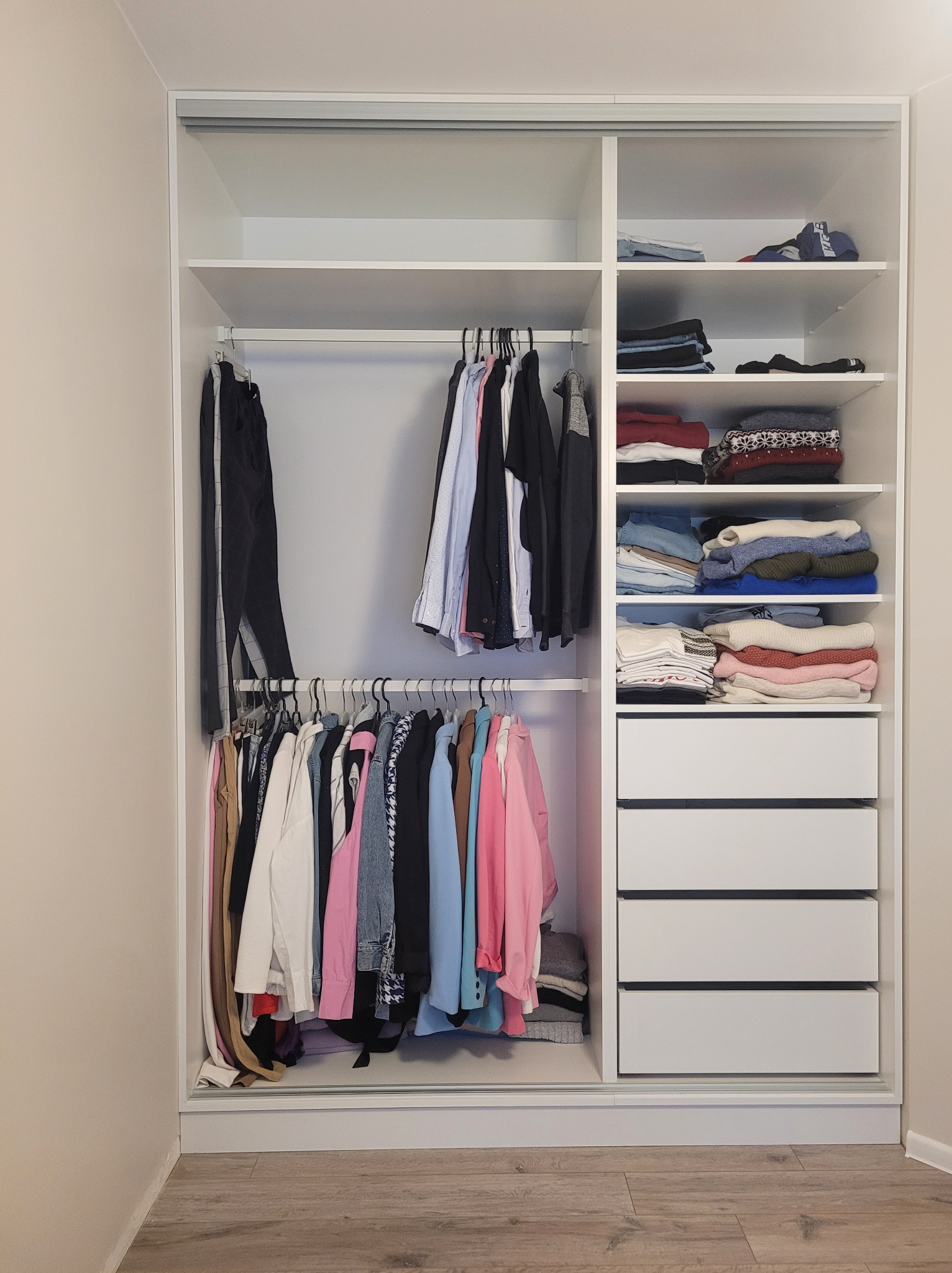 Biała szafa wnękowa z przesuwnymi drzwiami, wypełniona ubraniami na wieszakach, półkach i w szufladach. Widok wnętrza szafy.