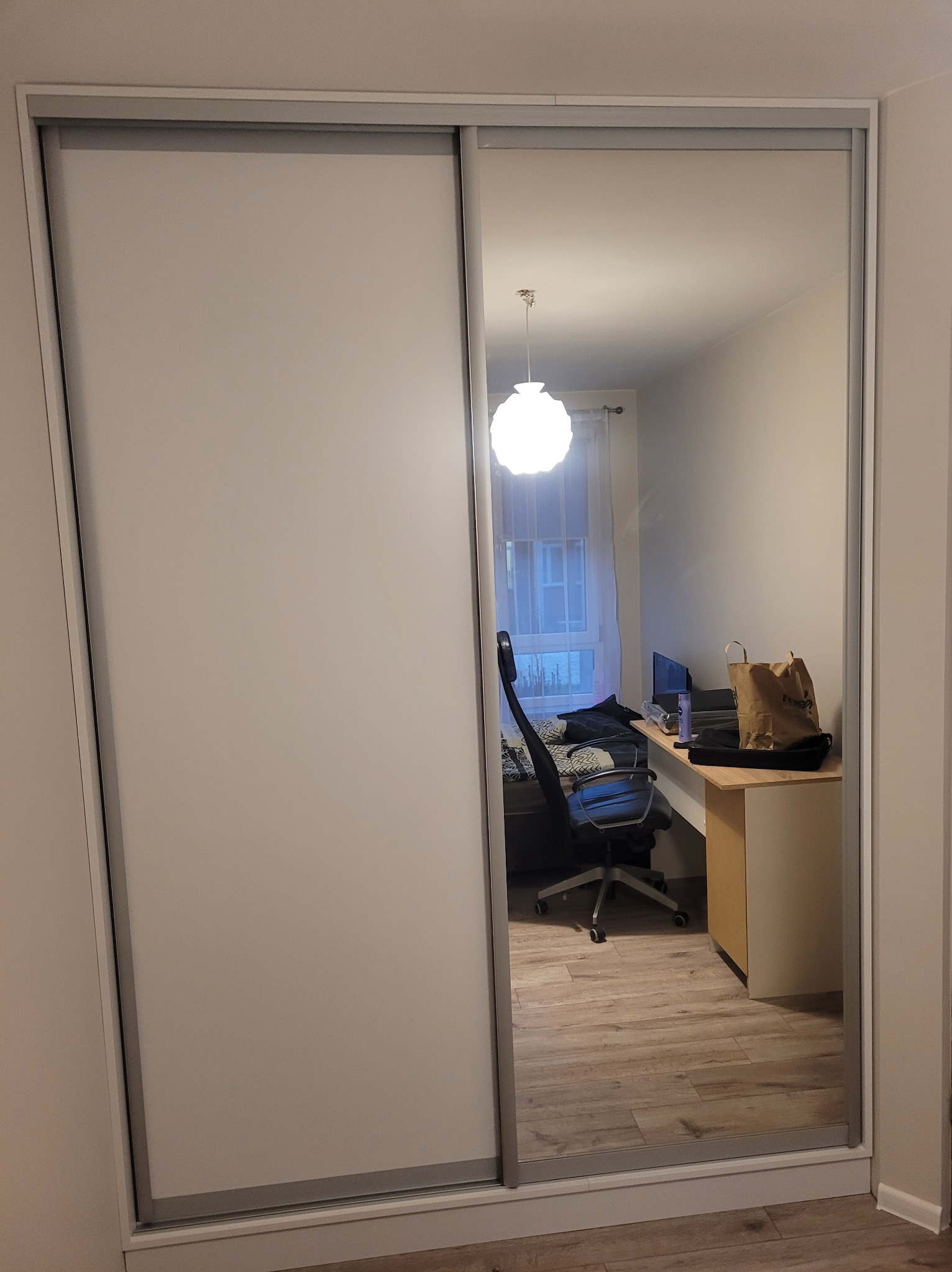 Biała szafa przesuwna z lustrem, odbijająca pokój z biurkiem, krzesłem i lampą. Widoczna część szafy z białymi drzwiami.