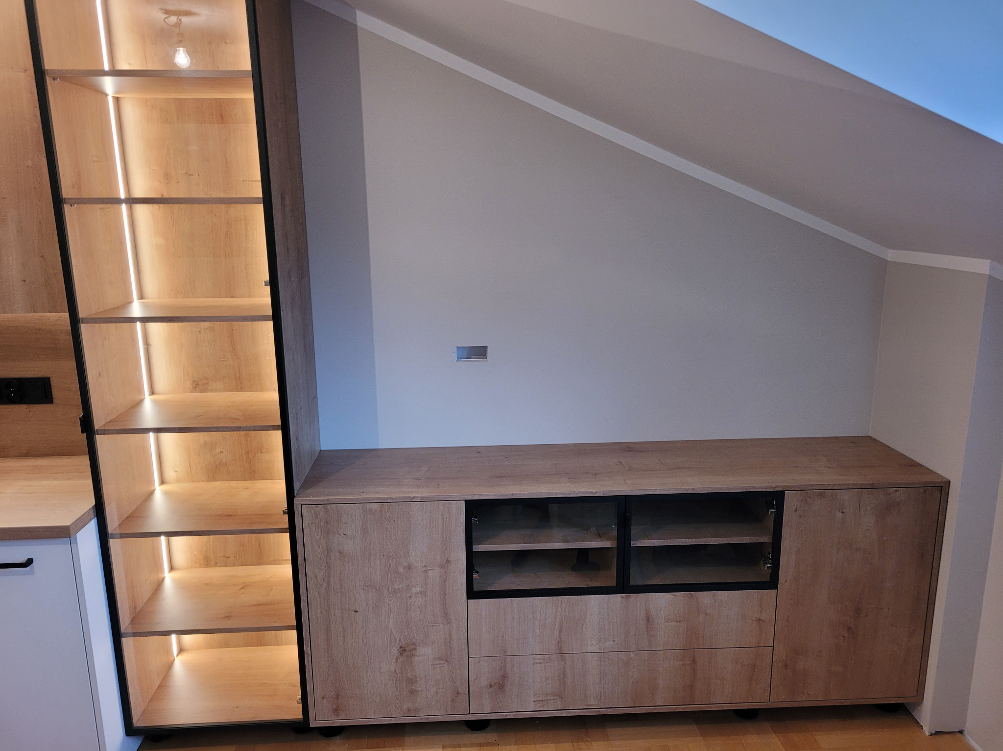 Drewniana zabudowa wnęki ze szklanymi drzwiczkami i podświetlanymi półkami, dopasowana do skosu dachu. Widoczna część kuchni z białymi frontami.