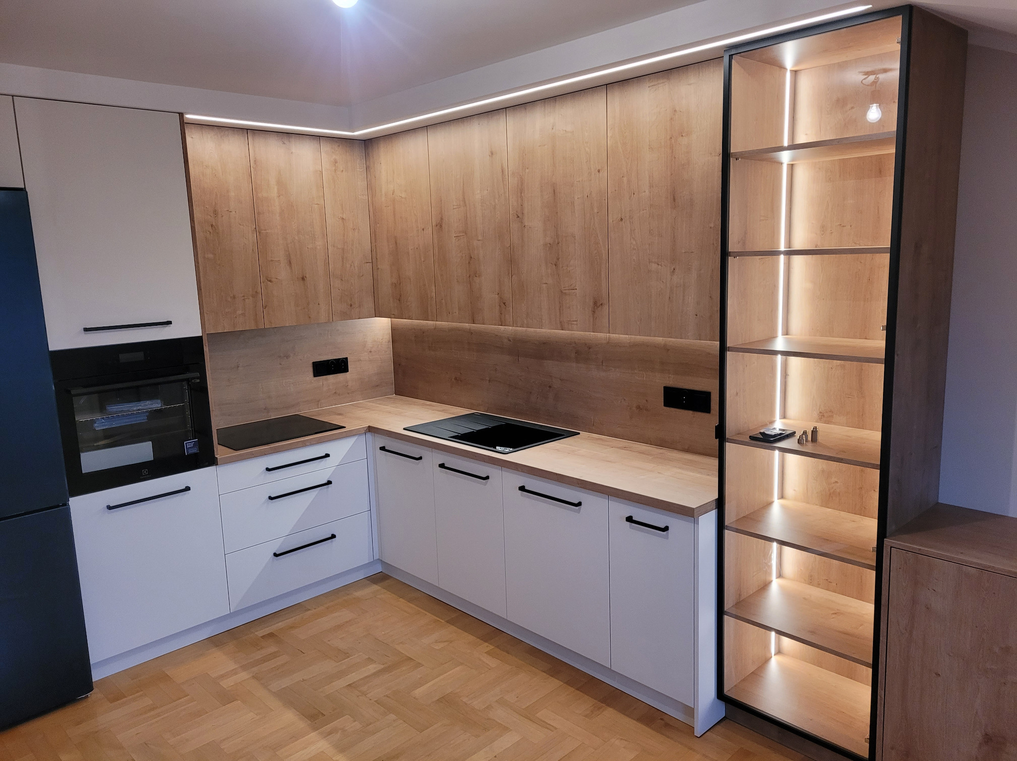 Nowoczesna kuchnia z białymi szafkami i drewnianymi frontami, oświetleniem LED pod szafkami wiszącymi i w otwartym regale z czarną ramą.