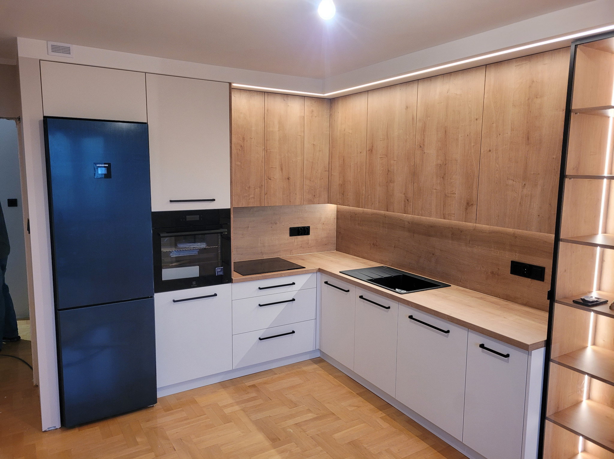 Nowoczesna kuchnia narożna z białymi frontami i drewnopodobnym blatem. Czarny sprzęt AGD i oświetlenie LED podkreślają minimalistyczny design.