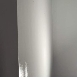 S.KOSS - Świeżo pomalowana, biała ściana z widocznymi śladami po niedoskonałościach szpachlowania pod światło, widoczne dwa małe otwory po gwoździach.