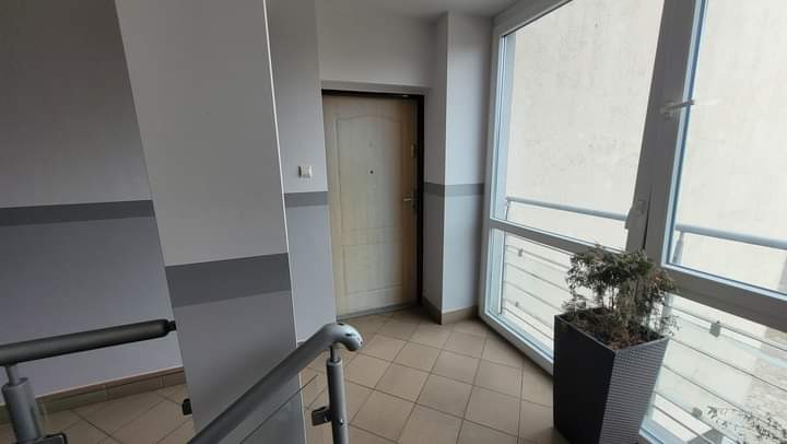 Klatka schodowa w bloku mieszkalnym po remoncie: świeżo pomalowane ściany w odcieniach szarości i bieli z dekoracyjnymi pasami, jasne płytki podłogowe, drzwi wejściowe do mieszkania i duże okno...