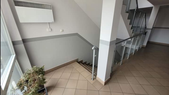 Klatka schodowa w biurowcu z szarymi ścianami, brązowymi płytkami na podłodze i metalowymi barierkami, na ścianie zamontowany grzejnik elektryczny.