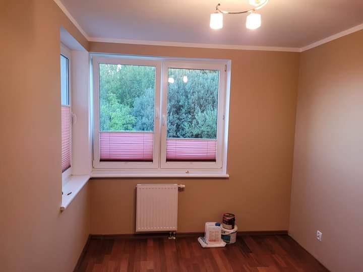 Pokój po odświeżeniu ścian w odcieniach beżu, widoczne puszki z farbą na podłodze przy grzejniku i oknach z żaluzjami.