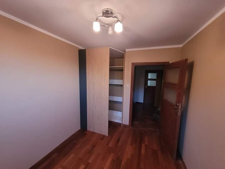 Pokój po remoncie z drewnianą podłogą, świeżo pomalowanymi ścianami w odcieniach beżu i otwartą szafą wnękową z półkami, widok na otwarte drzwi do kolejnego pomieszczenia.