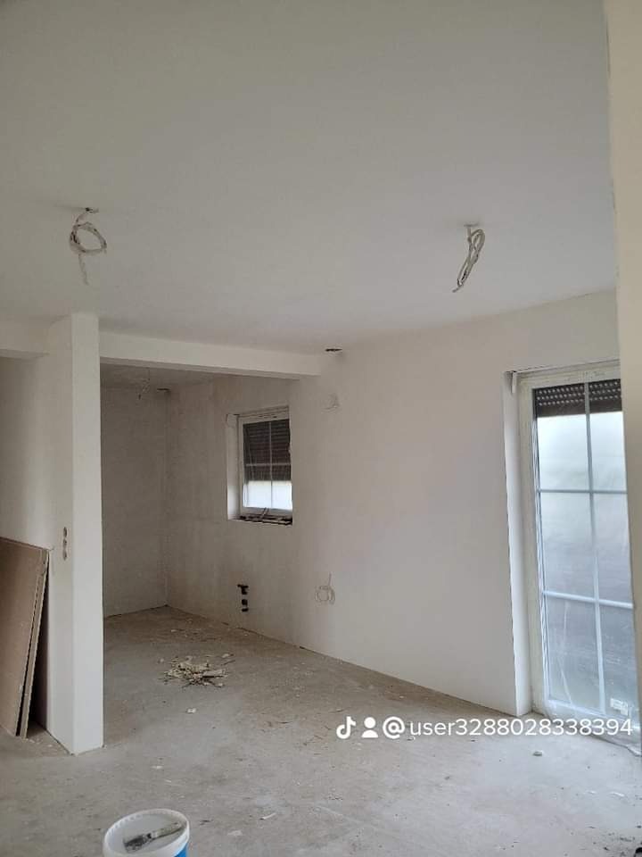 Wnętrze w trakcie remontu: świeżo wyszpachlowane ściany w jasnym odcieniu, widoczne kable wystające z sufitu, okno z żaluzjami i drzwi balkonowe z szybami, na podłodze resztki gruzu i wiadro po...