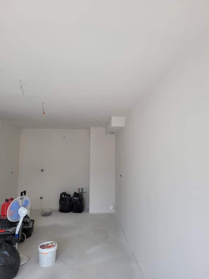 Wnętrze pokoju po świeżym pomalowaniu na biało, widoczne wiszące kable, wentylator i wiadro z farbą na podłodze.