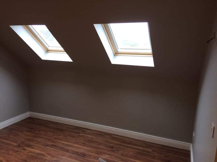 Pokój na poddaszu po remoncie, ściany pomalowane na szaro, drewniana podłoga, dwa okna dachowe z drewnianymi ramami.