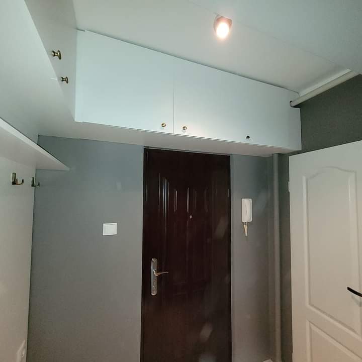 Przedpokój po remoncie: ciemne drzwi wejściowe, szara ściana, białe szafki pod sufitem i wieszaki na ubrania.