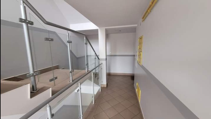 Klatka schodowa w budynku we Włocławku z szarymi ścianami, brązowymi płytkami na podłodze i szklaną balustradą. Ściany pomalowane w dwóch odcieniach szarości.