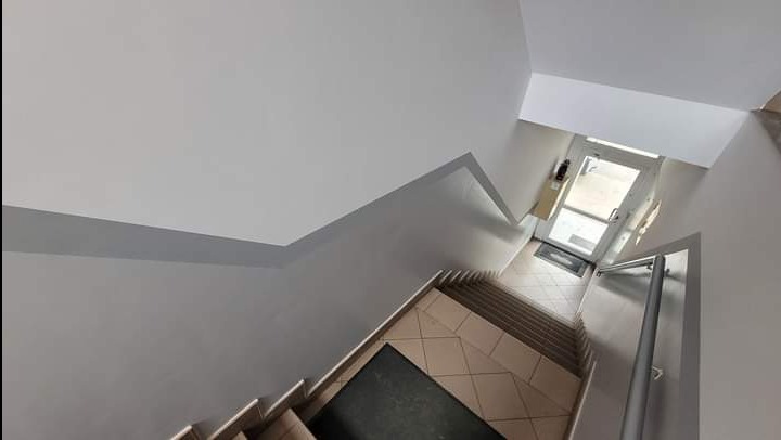 Klatka schodowa w budynku po odświeżeniu, ściany pomalowane na jasny szary kolor, widoczne schody z płytkami w odcieniach beżu i brązu, w tle przeszklone drzwi wejściowe.