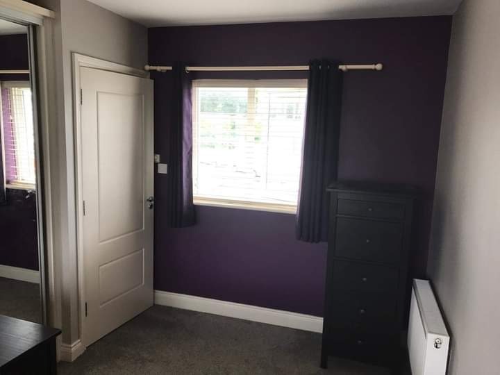 Pomieszczenie po odświeżeniu, ściana pomalowana na fioletowo, widoczne białe drzwi, okno z żaluzjami, czarna komoda i grzejnik.