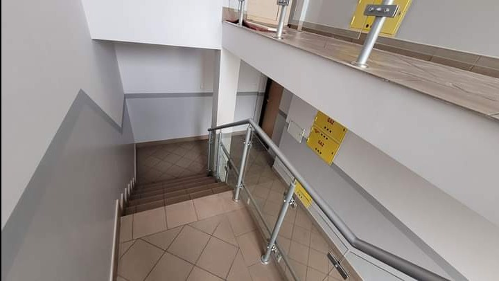 Klatka schodowa w budynku biurowym po remoncie, widoczne świeżo pomalowane ściany z szarym pasem dekoracyjnym u dołu, płytki podłogowe w beżowym odcieniu i szklane balustrady.