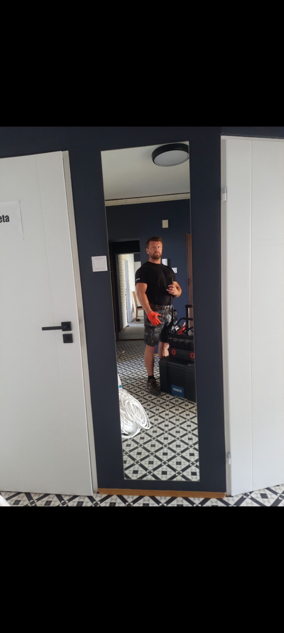 Autoportret mężczyzny w czarnej koszulce, krótkich spodenkach moro i pomarańczowych rękawicach roboczych, stojącego przed lustrem w trakcie pracy, obok skrzynka z narzędziami i zwój kabli, widoczna...