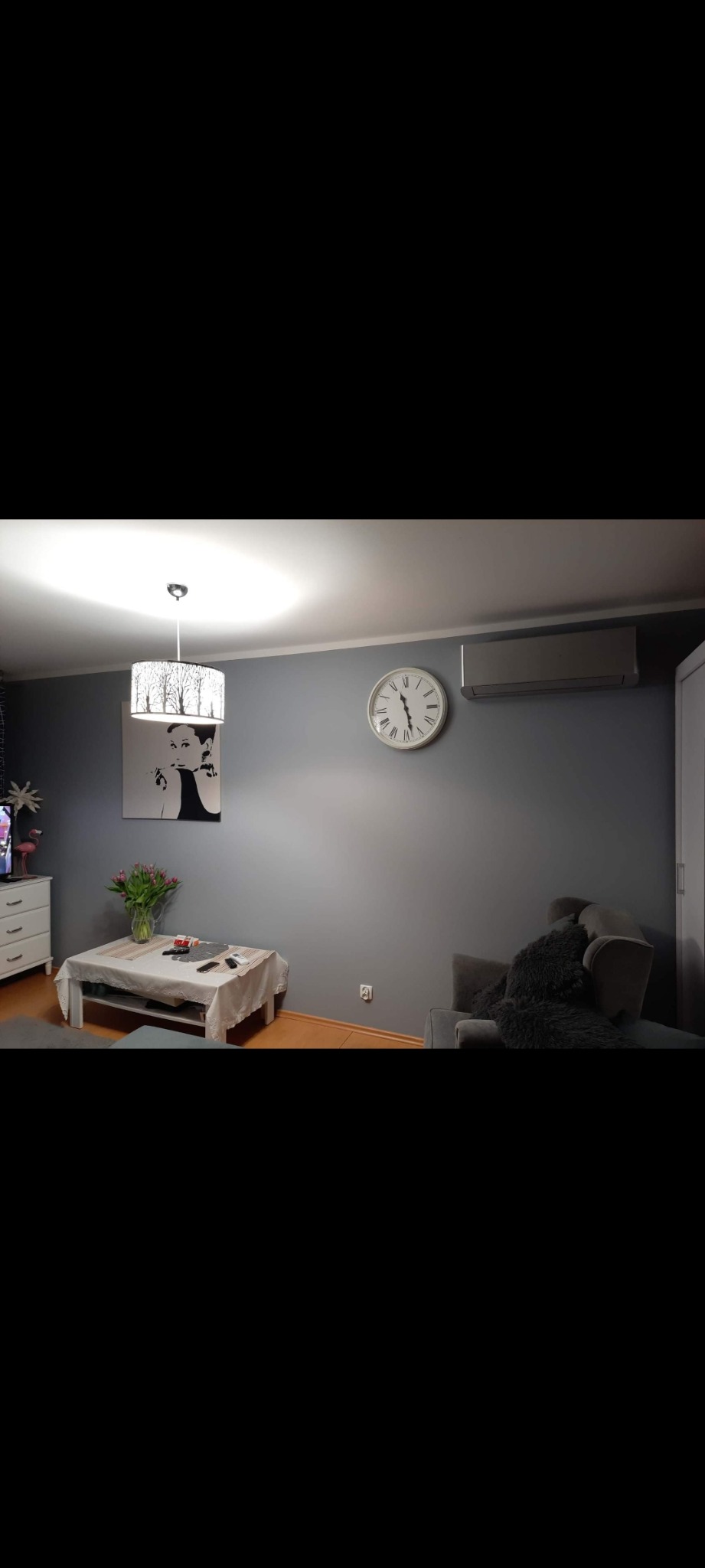 Szare wnętrze pokoju z klimatyzatorem na ścianie, zegarem w stylu retro i obrazem Audrey Hepburn.