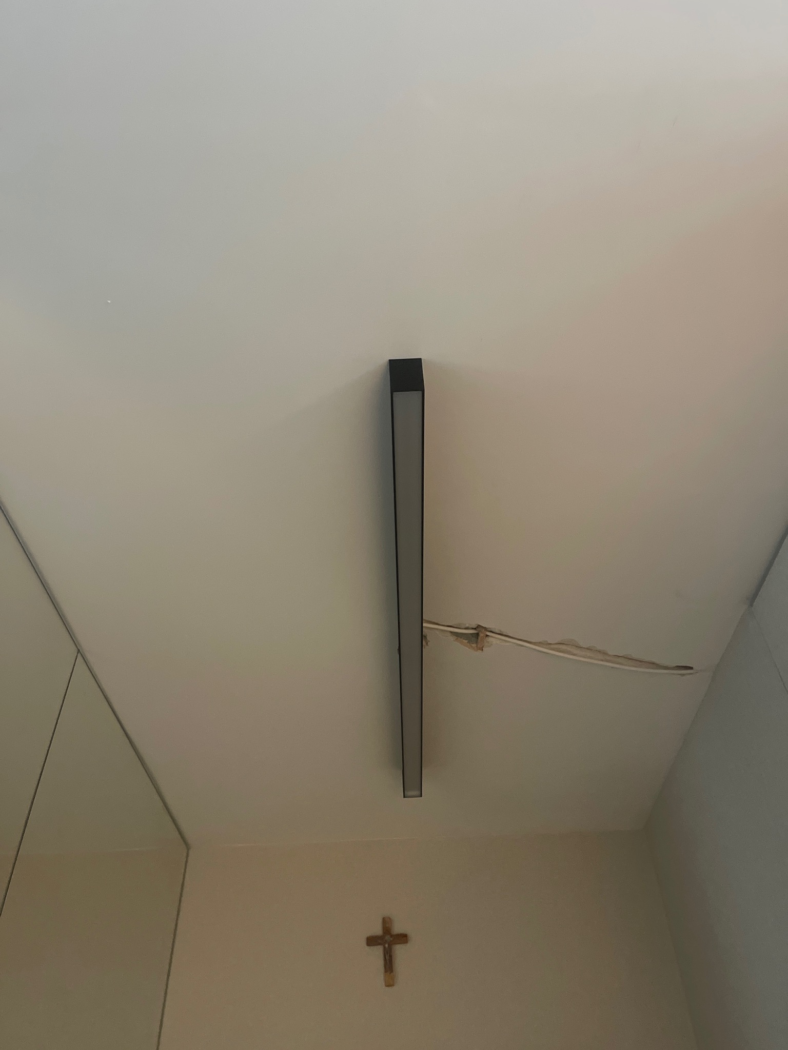 Czarna lampa LED zamontowana na suficie z widocznymi kablami wystającymi ze ściany, poniżej krzyż na ścianie.