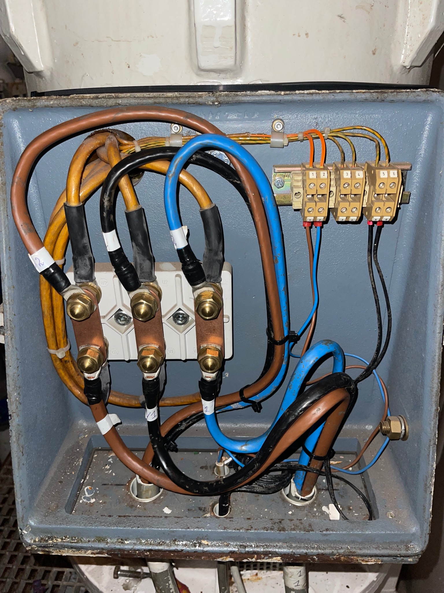 Otwarta, szara skrzynka elektryczna z widocznymi, podłączonymi kablami o różnych kolorach (brązowy, żółty, niebieski, czarny) i złączkami, niektóre kable oznaczone białymi etykietami.