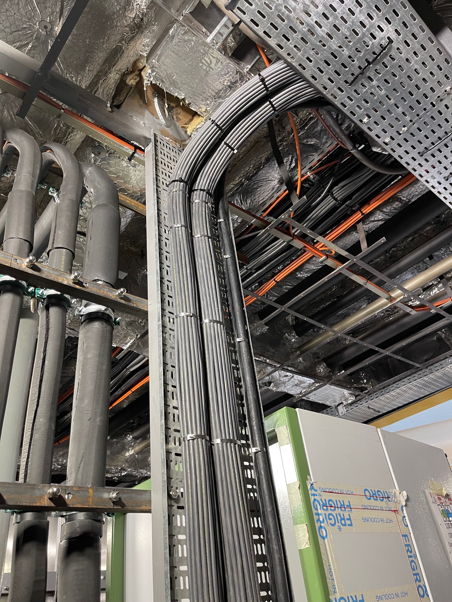 Widok instalacji przemysłowej z licznymi przewodami ułożonymi w korytach kablowych pod sufitem, obok rur izolowanych termicznie, z widocznymi elementami konstrukcyjnymi i fragmentami urządzeń...