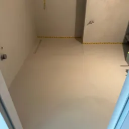 Jasnoszara, gładka powierzchnia w pomieszczeniu z taśmą maskującą na ścianach, widoczne elementy instalacji na suficie, ujęcie z dołu.