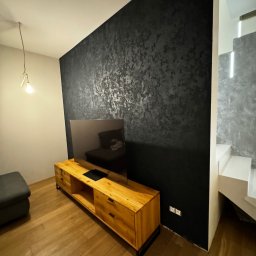 DecorativeGroup - Ściana w salonie wykończona ciemnym mikrocementem ze srebrnymi refleksami, drewniana szafka RTV i podłoga z parkietu, widoczne schody z podświetleniem LED.