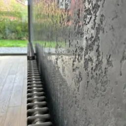 Szara ściana z efektem betonu architektonicznego, widoczny grzejnik żeberkowy i okno z widokiem na zieleń ogrodu.