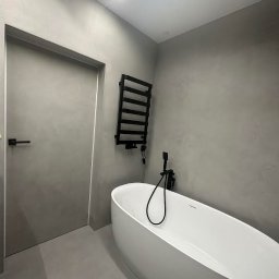 DecorativeGroup - Minimalistyczna łazienka w odcieniach szarości z wolnostojącą białą wanną, czarną armaturą i grzejnikiem drabinkowym, wykończona gładką, jednolitą powierzchnią ścian i podłogi.