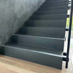 DecorativeGroup - Nowoczesne schody wewnętrzne z szarego materiału, kontrastujące z drewnianą podłogą i ścianą z szarego kamienia, widoczne przez okno zielone krzewy.