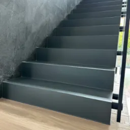 Nowoczesne schody wewnętrzne z szarego materiału, kontrastujące z drewnianą podłogą i ścianą z szarego kamienia, widoczne przez okno zielone krzewy.