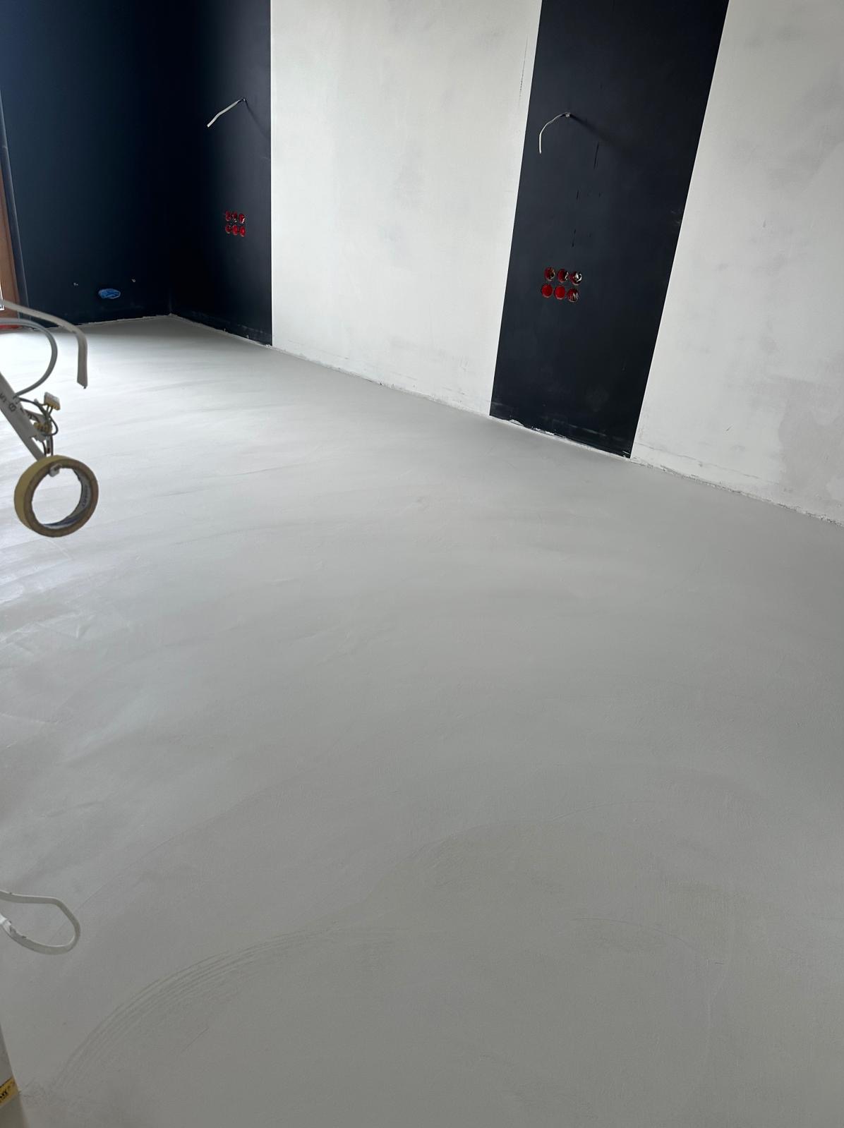Świeżo wylana, jasnoszara posadzka w pomieszczeniu z czarno-białymi akcentami na ścianach, widoczny fragment narzędzia z taśmą malarską.