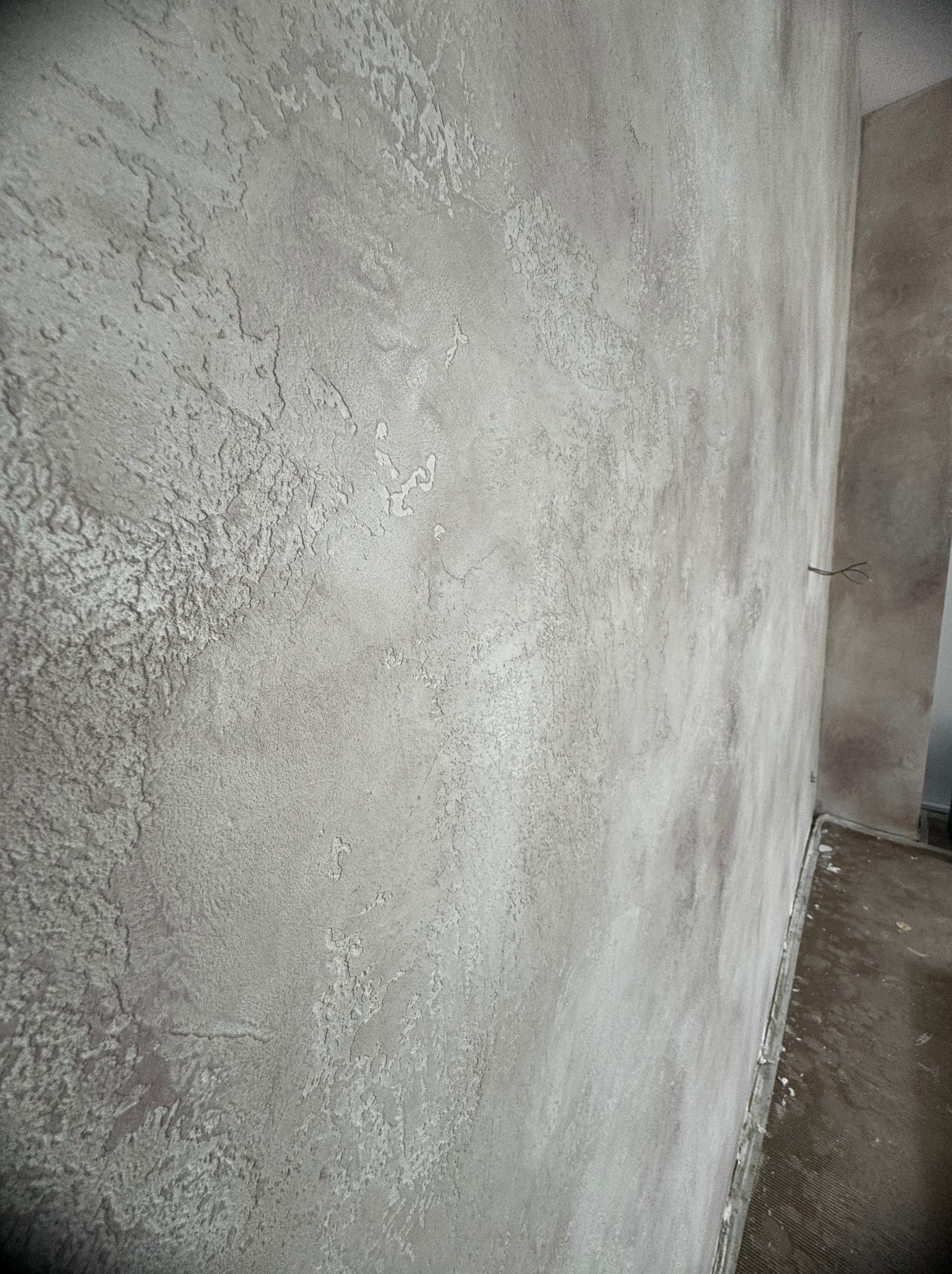 Ściana pokryta szarą, dekoracyjną strukturą tynkową z widoczną fakturą i nierównościami, oświetlona naturalnym światłem, z fragmentem podłogi i wystającymi kablami w rogu.