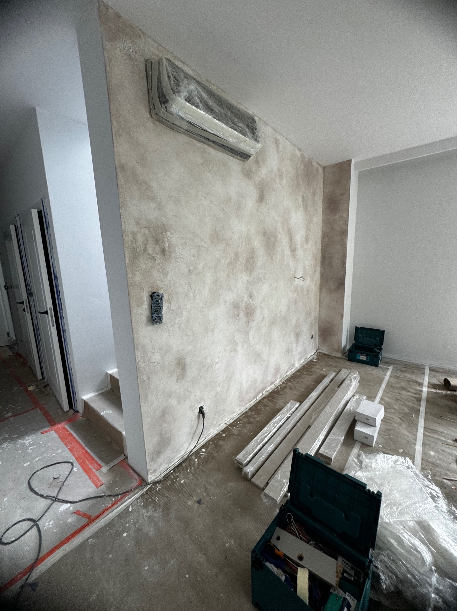 Ściana z dekoracyjną, beżowo-brązową fakturą, zabezpieczony folią klimatyzator, widoczne puszki elektryczne, listwy przypodłogowe w folii, skrzynka z narzędziami oraz fragment schodów w trakcie...