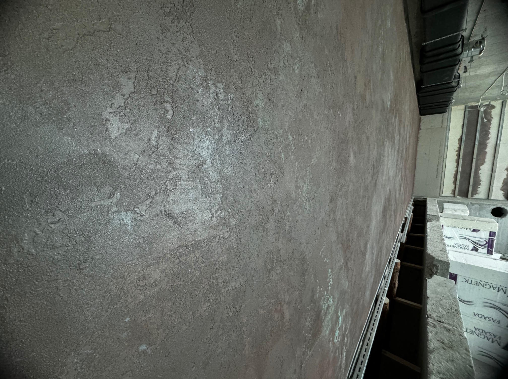 Szara ściana z wyraźną, nieregularną fakturą, widoczna pod kątem, z fragmentem sufitu z instalacją wentylacyjną i elementami konstrukcyjnymi w tle.