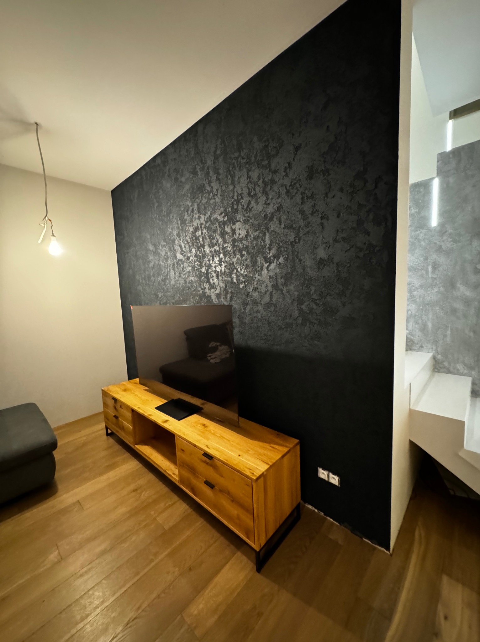 Ściana w salonie wykończona ciemnym mikrocementem ze srebrnymi refleksami, drewniana szafka RTV i podłoga z parkietu, widoczne schody z podświetleniem LED.