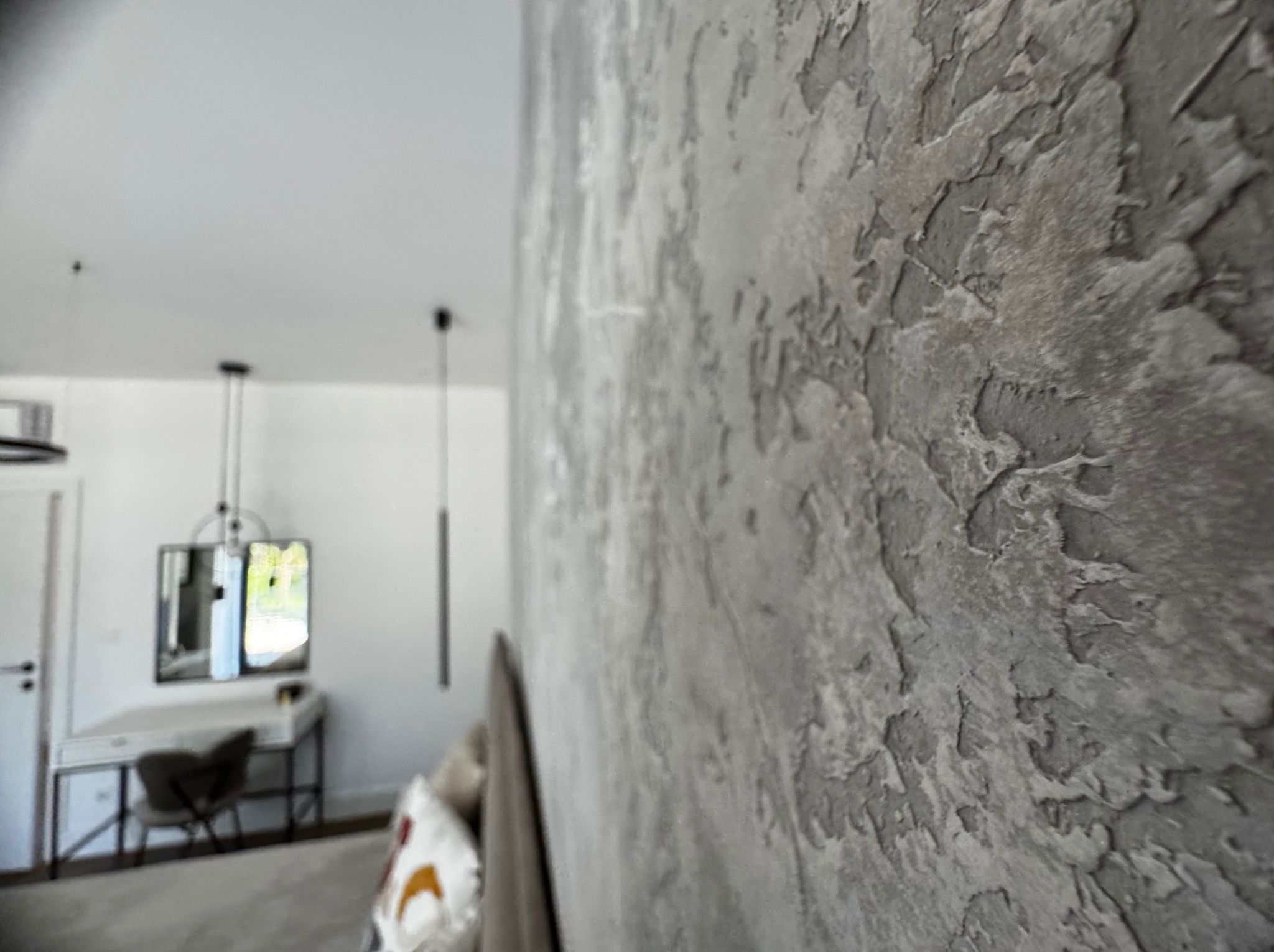 Szara ściana z wyraźną, nieregularną fakturą, przypominającą kamień, w tle rozmyte wnętrze sypialni z toaletką i lampami wiszącymi.