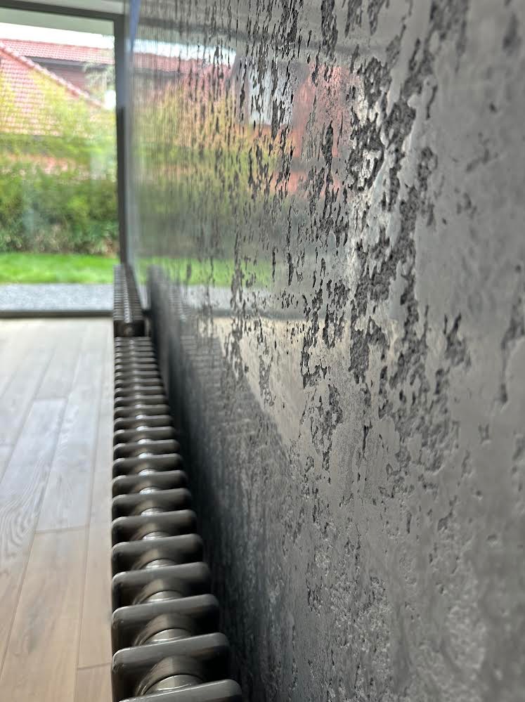 Szara ściana z efektem betonu architektonicznego, widoczny grzejnik żeberkowy i okno z widokiem na zieleń ogrodu.