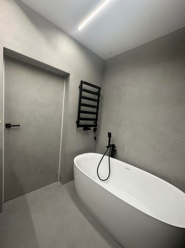 Minimalistyczna łazienka w odcieniach szarości z wolnostojącą białą wanną, czarną armaturą i grzejnikiem drabinkowym, wykończona gładką, jednolitą powierzchnią ścian i podłogi.
