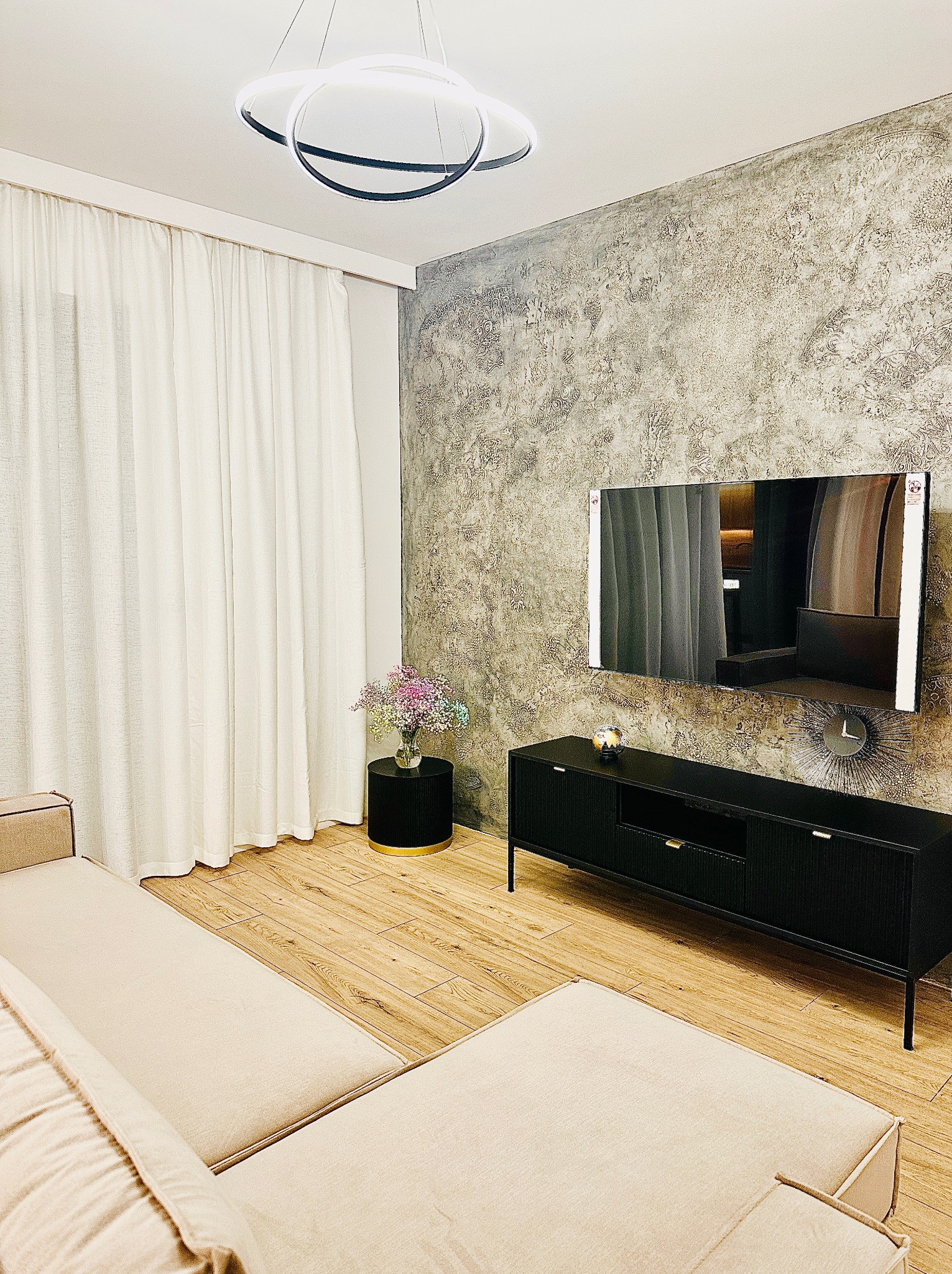 Salon z dekoracyjną ścianą o fakturze betonu, drewnianą podłogą, beżową sofą i czarną szafką RTV pod telewizorem, oświetlony nowoczesną lampą wiszącą.