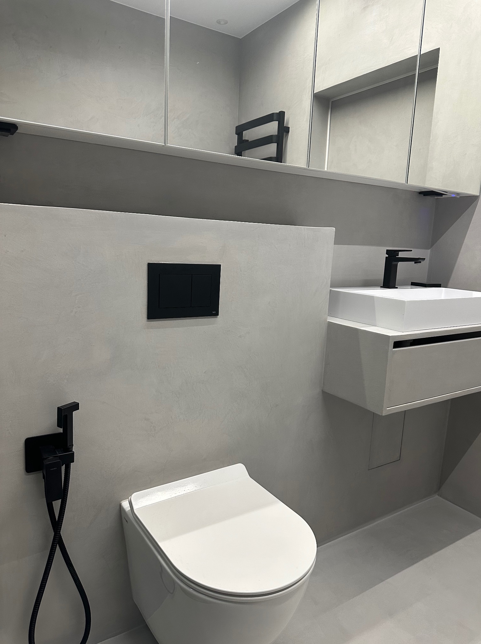 Minimalistyczna łazienka w odcieniach szarości z białą ceramiką, czarną armaturą i bidetką natryskową, wykończona gładzią imitującą beton.