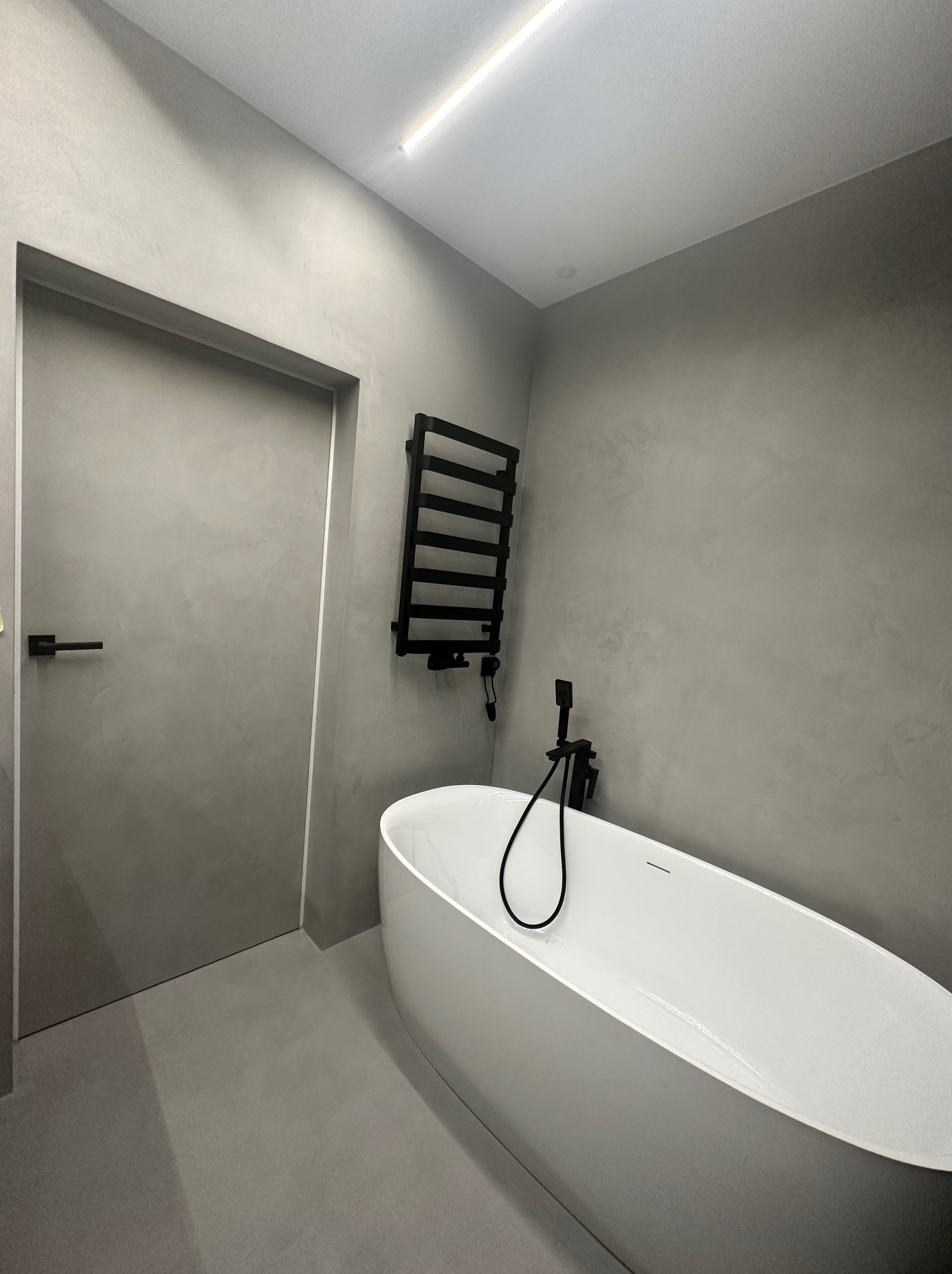 Minimalistyczna łazienka w szarościach z wolnostojącą białą wanną, czarną armaturą i grzejnikiem drabinkowym, wykończona tynkiem dekoracyjnym imitującym beton.