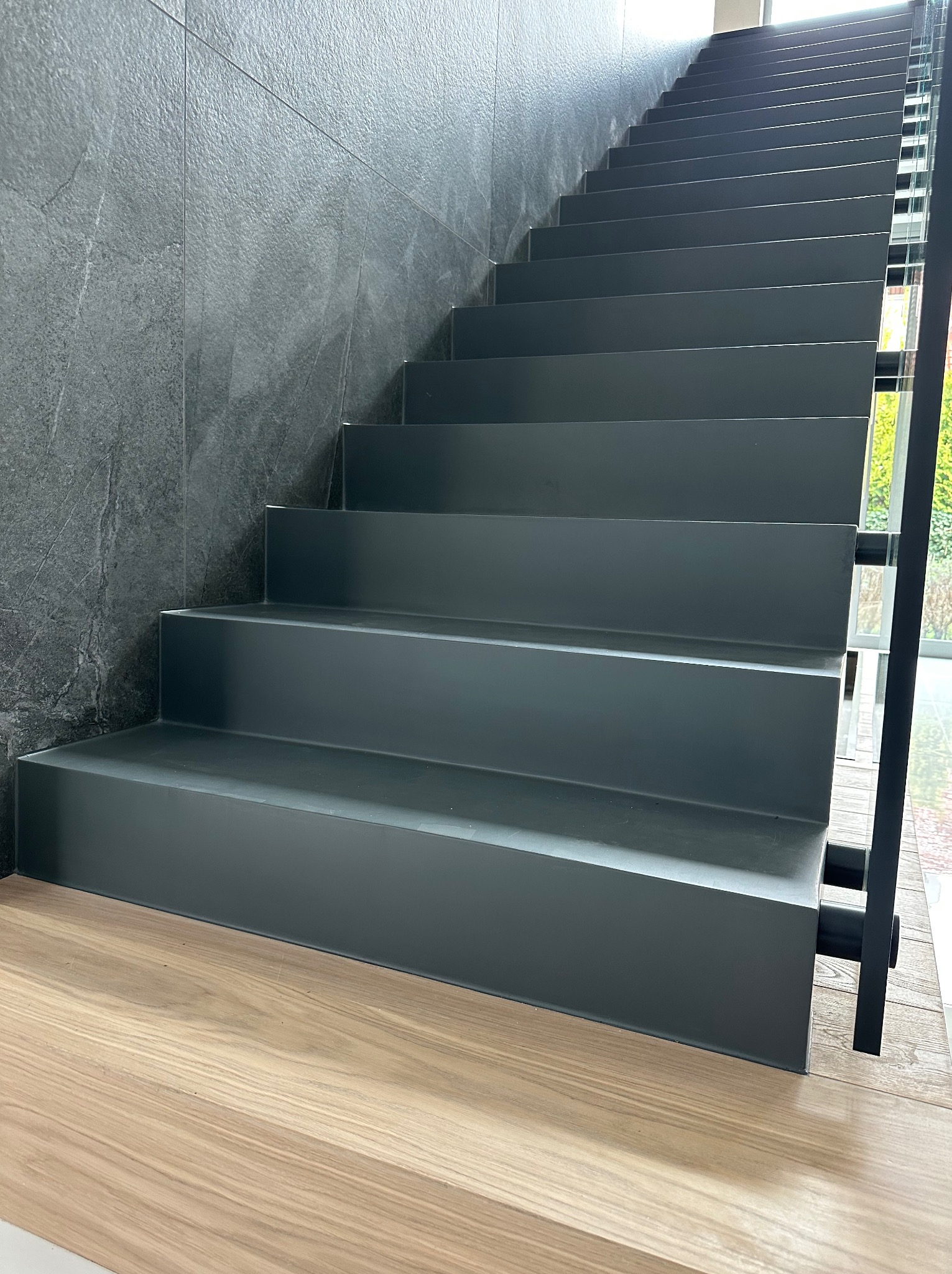 Nowoczesne schody wewnętrzne z szarego materiału, kontrastujące z drewnianą podłogą i ścianą z szarego kamienia, widoczne przez okno zielone krzewy.