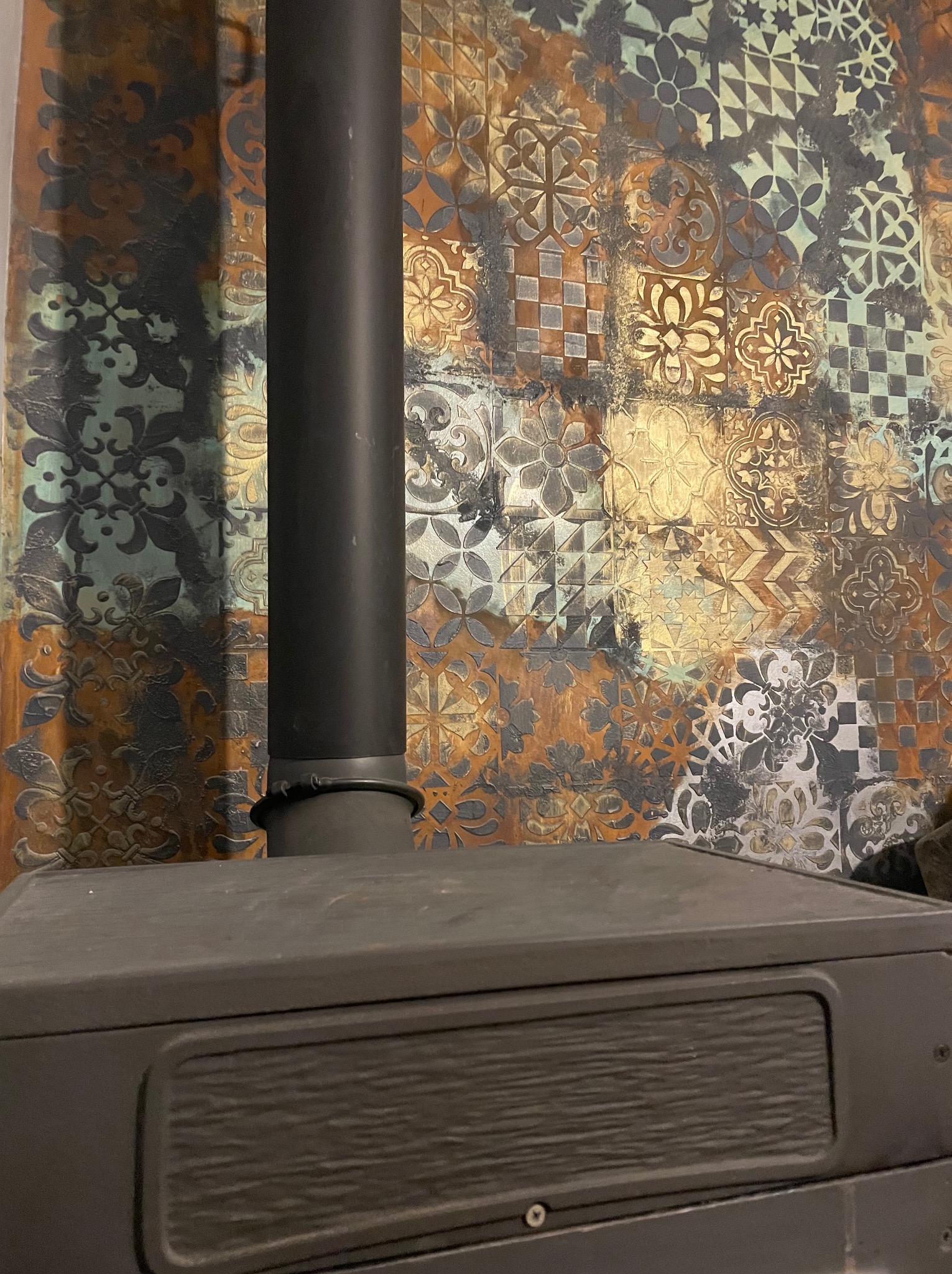 Fragment ściany z dekoracyjnymi panelami imitującymi patchwork płytek w odcieniach brązu, złota, szarości i turkusu, z widocznym piecem i czarną rurą kominową.
