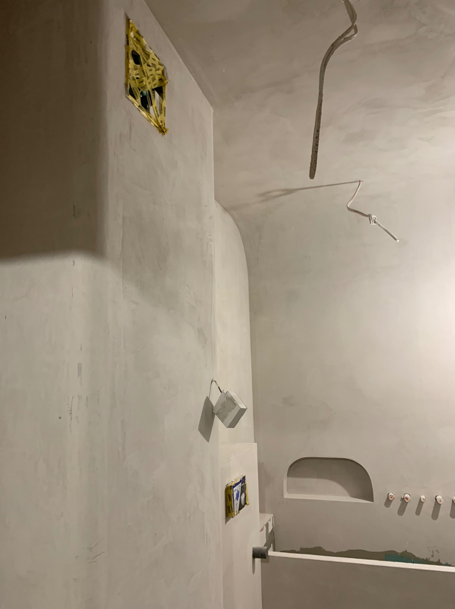 Wnętrze łazienki w trakcie prac wykończeniowych, ściany świeżo pokryte tynkiem, widoczne zabezpieczone otwory instalacyjne i wystające przewody elektryczne.