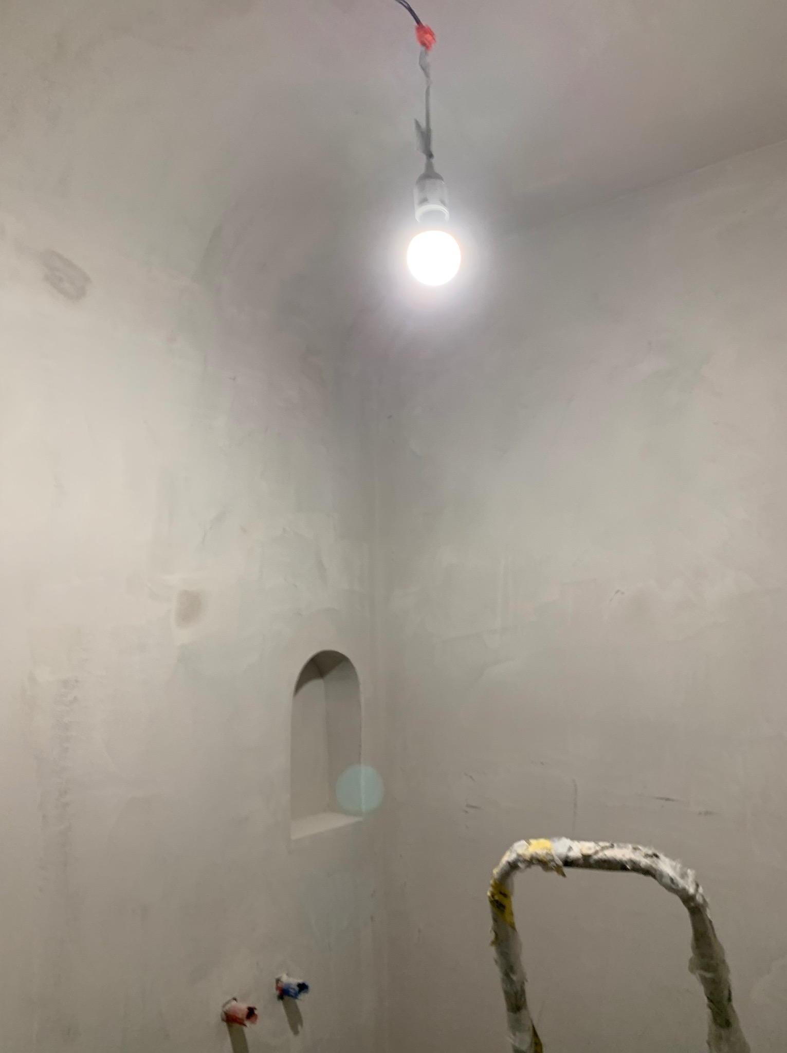 Wnętrze łazienki w trakcie tynkowania, widoczna surowa powierzchnia ścian z wnęką, prowizoryczne oświetlenie żarówką i fragment drabiny malarskiej.
