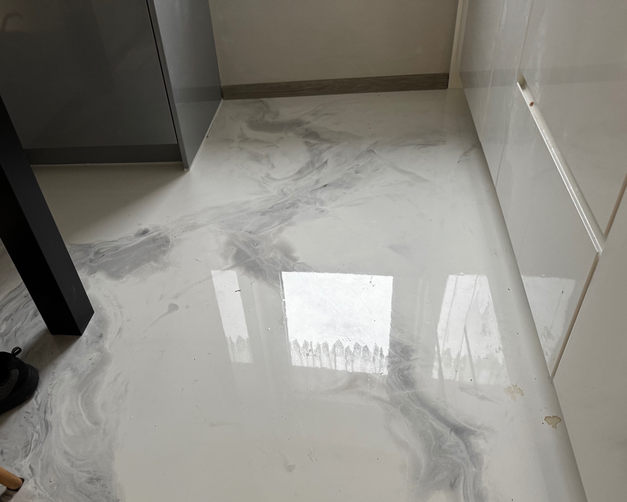 Biała, błyszcząca podłoga z szarymi marmurkowymi wzorami, odbijająca światło w pomieszczeniu z białymi i szarymi meblami, widoczny fragment buta i nogi stołu.