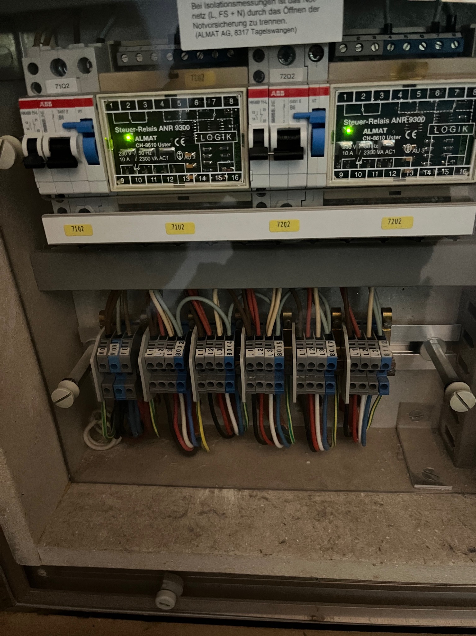 Otwarta skrzynka elektryczna z widocznymi przewodami podłączonymi do listew zaciskowych oraz modułami sterującymi Almat ANR 9300, z etykietami 71Q2 i 72Q2.